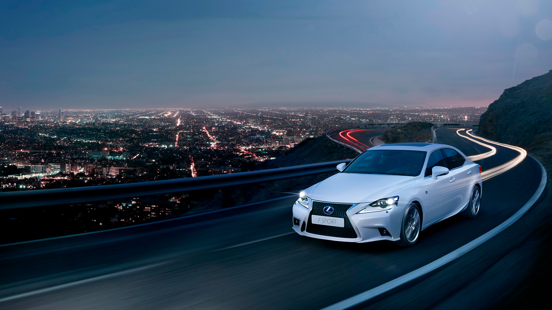 Vista frontal del Lexus IS 300h conduciendo de noche con una ciudad de fondo