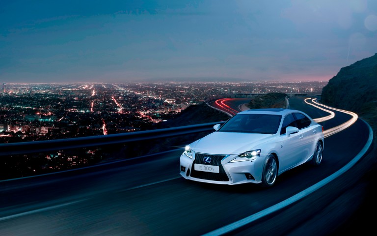 Vista frontal del Lexus IS 300h conduciendo de noche con una ciudad de fondo
